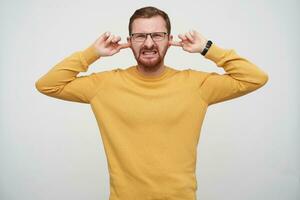 mécontent Jeune jolie barbu brunette homme dans des lunettes ayant court la Coupe de cheveux, fermeture le sien yeux et renfrogné visage tandis que éviter bruyant des sons, isolé plus de blanc Contexte photo