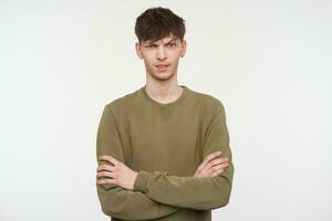 adolescent gars, homme avec brunette cheveux, perçant et poil. portant kaki Couleur chandail. à la recherche incrédule, en portant bras franchi. en train de regarder à le caméra isolé plus de blanc Contexte photo