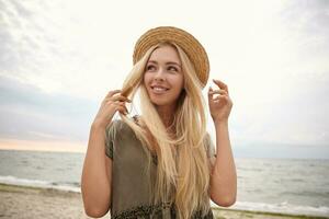 portrait de attrayant Jeune blond femme avec longue cheveux à la recherche de côté avec charmant sourire, posant plus de bord de mer sur couvert dans décontractée robe et canotier chapeau photo