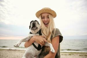 marrant coup de magnifique Jeune femme avec longue blond cheveux portant décontractée vêtements, en marchant le long de plage sur couvert avec sa chien, à la recherche à caméra joyeusement et souriant largement photo