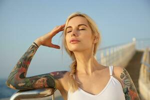 Extérieur portrait de jolie Jeune blond femelle avec tatouages portant sa cheveux dans tresser, posant plus de front de mer avec fermé yeux, exposer sa visage à Soleil photo
