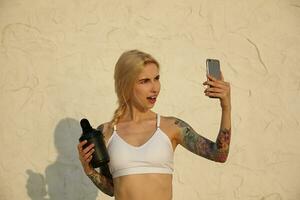 Extérieur coup de jolie Jeune blond femelle avec tatouages et décontractée coiffure permanent plus de blanc mur sur chaud ensoleillé jour, en portant noir Mixeur et fabrication selfie avec téléphone intelligent photo