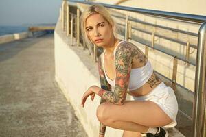 attrayant tatoué Jeune blond femelle avec décontractée coiffure posant plus de bord de mer promenade sur de bonne heure été matin, portant sportif vêtements et baskets, à la recherche de côté pensivement photo