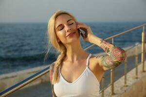 Jeune attrayant tatoué blond femme avec décontractée coiffure posant plus de bord de mer voir, portant blanc sportif haut, fabrication appel avec sa téléphone intelligent et en gardant yeux fermé photo