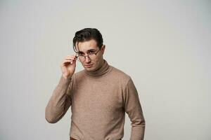 studio photo de jolie Jeune brunette Masculin avec branché coiffure en gardant élevé main sur le sien lunettes tandis que à la recherche à caméra, permanent plus de blanc Contexte dans à la mode vêtements