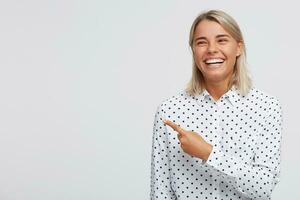 portrait de de bonne humeur charmant blond Jeune femme porte polka point chemise en riant et montrer du doigt à le côté par doigt isolé plus de blanc Contexte photo