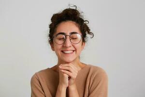 portrait de souriant géorgien Jeune femme avec fermé yeux porte beige arrêtez-vous et des lunettes garde mains dans prier position et fabrication une souhait isolé plus de blanc Contexte photo