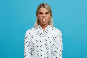 fermer de amusant espiègle blond Jeune femme porte polka point chemise fait du marrant visage et spectacles langue isolé plus de bleu Contexte photo