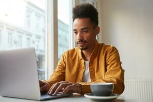 Jeune africain américain homme les usages moderne les technologies, est assis dans de face de portable ordinateur, porte Jaune chemise, les boissons aromatique café dans cafétéria, crée Nouveau contenu pour le sien Blog. photo