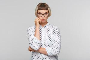 portrait de réfléchi jolie blond Jeune femme porte polka point chemise et des lunettes regards concentré et garde mains plié isolé plus de blanc Contexte photo