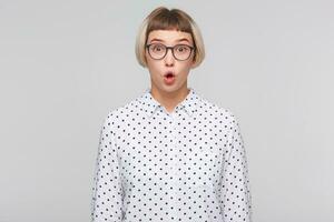 fermer de surpris mignonne blond Jeune femme porte polka point chemise et lunettes regards amusant et étonné isolé plus de blanc Contexte photo