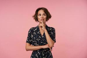 positif Jeune brunette Dame avec court la Coupe de cheveux en portant index sur sa bouche, demander à garder secret, portant noir à motifs robe tandis que posant plus de rose Contexte photo