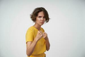 sévère Jeune brunette Dame avec court la Coupe de cheveux élevage les poings et étant prêt à défendre se, à la recherche menaçant à caméra et renfrogné les sourcils, posant plus de blanc Contexte photo