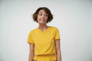 portrait de embarrassé Jeune femelle Dame avec court marron cheveux à la recherche de côté et montrant les dents confusément, en gardant mains le long de sa corps tandis que posant plus de blanc Contexte photo