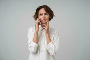 sérieux à la recherche Jeune brunette femme avec court la Coupe de cheveux en portant mobile téléphone et ayant conversation avec sa les partenaires, renfrogné les sourcils d'un air songeur, isolé plus de blanc Contexte photo