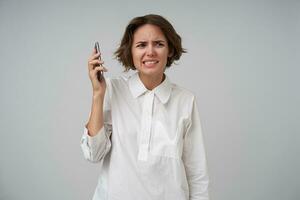 intérieur coup de mécontent Jeune Dame avec court marron cheveux ayant méchant conversation sur téléphone, à la recherche de côté avec moue et torsion sa bouche, permanent plus de blanc Contexte dans formel vêtements photo