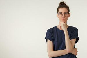 portrait de attrayant Jeune femme dans des lunettes regards en haut réfléchit une pensée ou idée, réfléchi, garde poing près le menton, porte décontractée T-shirt, sur une blanc Contexte photo