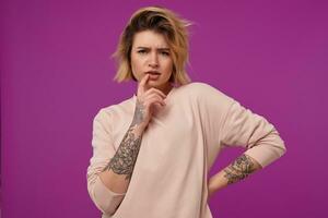 intérieur portrait de Jeune teint blond femme, avec tatoué bras émouvant sa lèvres avec une doigt, réfléchi faciale expression, isolé plus de violet Contexte photo