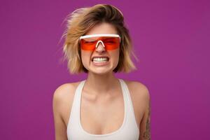 intérieur portrait de Jeune teint blond femme, avec tatoué bras porte Orange élégant lunettes, pressage sa les dents avec en colère faciale expression. isolé plus de violet Contexte photo