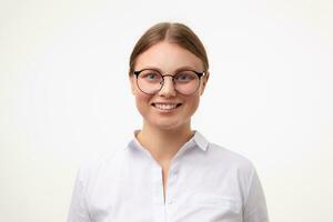 portrait de Jeune aux yeux bleus blond femelle dans des lunettes montrant sa parfait blanc les dents tandis que à la recherche volontier à caméra, permanent plus de blanc Contexte dans blanc chemise photo