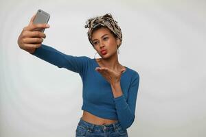 intérieur photo de Jeune jolie foncé aux cheveux frisé femme avec recueillies frisé cheveux en gardant sa main élevé tandis que fabrication selfie sur téléphone intelligent et pliant lèvres dans air baiser, isolé plus de blanc Contexte