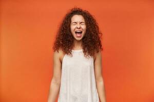 adolescent fille, malheureux à la recherche femme avec frisé gingembre cheveux. portant blanc épaules dénudées chemisier. en criant louange avec fermé yeux. hurler dans colère. supporter isolé plus de Orange Contexte photo