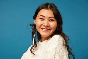 intérieur photo de Jeune cheveux bruns asiatique femelle avec boucles étant dans agréable ambiance tandis que écoute à musique, habillé dans blanc tricoté chandail tandis que posant plus de bleu Contexte