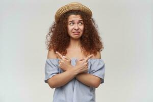 portrait de attractif, adulte roux fille avec frisé cheveux. portant rayé épaules dénudées chemisier et chapeau. en train de regarder à le droite à copie espace et montrer du doigt tous les deux côtés, isolé plus de Orange Contexte photo