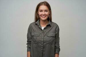 Jeune dame, jolie femme avec marron longue cheveux. portant gris chemise et souriant. introduction sa soi. sur de soi posture. en train de regarder à le caméra isolé plus de gris Contexte photo