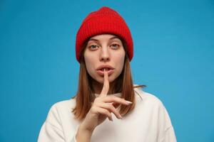 adolescent fille, femme avec brunette cheveux. portant blanc chandail et rouge chapeau. montrant silence signe, garder assez. gens concept. fermer, en train de regarder à le caméra, isolé plus de bleu Contexte photo