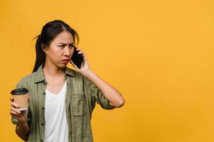 une jeune femme asiatique parle par téléphone et tient une tasse de café avec une expression négative, des cris excités, pleure émotionnellement en colère dans un tissu décontracté et se tient isolée sur fond jaune. concept d'expression faciale.