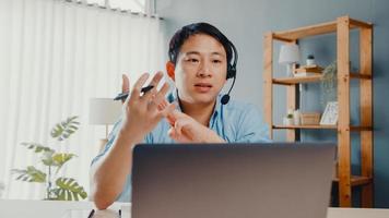 un jeune homme d'affaires asiatique porte des écouteurs à l'aide d'un ordinateur portable et parle à ses collègues du plan lors d'un appel vidéo pendant qu'il travaille à domicile dans le salon. auto-isolement, distanciation sociale, quarantaine pour la prévention du covid.