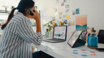 une jeune femme d'affaires asiatique appelle un téléphone portable avec des collègues de l'entreprise à propos du plan de marché du graphique des finances du travail dans un ordinateur portable et une tablette à la maison. une étudiante apprend en ligne, travaille à domicile.