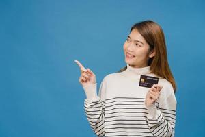 jeune femme asiatique montre une carte bancaire de crédit avec une expression positive, sourit largement, vêtue de vêtements décontractés, se sentant heureuse et isolée sur fond bleu. concept d'expression faciale. photo