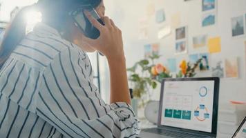 une jeune femme d'affaires asiatique appelle un téléphone portable avec des collègues de l'entreprise à propos du plan de marché du graphique des finances du travail dans un ordinateur portable et une tablette à la maison. une étudiante apprend en ligne, travaille à domicile.