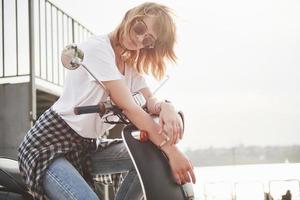 portrait d'une belle fille hipster assise sur un scooter rétro noir, souriant posant et profitant du chaud soleil du printemps. photo