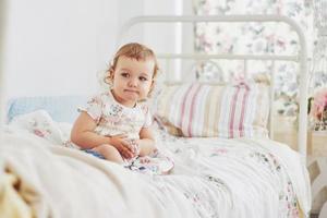 notion d'enfance. petite fille en robe mignonne implantation au lit jouant avec des jouets à la maison. chambre d'enfant vintage blanche photo