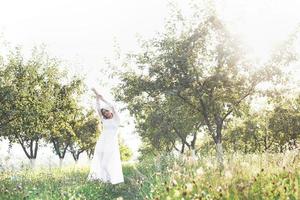 une jeune fille vêtue d'une longue robe blanche se promène dans le jardin. beau coucher de soleil à travers les feuilles des arbres photo