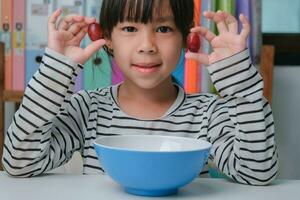 peu fille en mangeant Frais rouge les raisins à Accueil dans le vivant chambre. mignonne Jeune asiatique fille mange en bonne santé des fruits et Lait pour sa repas. en bonne santé nourriture dans enfance photo