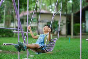 content asiatique fille ayant amusement en jouant dans le terrain de jeux pendant été. mignonne peu fille balançant dans le terrain de jeux avec une sourire et rire. actif été loisir pour enfants. photo