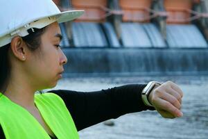 femelle ingénieur dans blanc chapeau à la recherche à montre intelligente tandis que travail à l'extérieur à barrage avec hydro-électrique Puissance plante et irrigation. photo