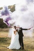 la mariée et jeune marié en jouant avec coloré fumée dans violet mains, câlins et bisous. fumée bombes à une mariage. photo