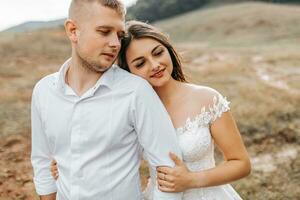 une élégant couple de européen jeunes mariés. une souriant la mariée dans une blanc robe câlins le jeune marié de derrière. le jeune marié est habillé dans une blanc chemise, sans pour autant une cravate. Extérieur mariage dans le montagnes photo