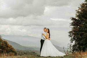 une mariage couple tendrement câlins dans le montagnes. le jeune marié câlins le la mariée par le épaules. une magnifique et magnifique de la mariée robe. photo