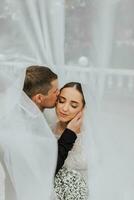 une élégant couple de européen jeunes mariés. souriant la mariée dans une blanc robe. le marié, habillé dans une classique noir costume, blanc chemise, baisers le la mariée sur le temple en dessous de le voile. mariage dans la nature photo