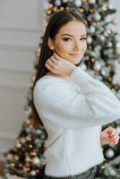 une magnifique et élégant Jeune fille dans une blanc chandail pose dans le Noël lumières sur le Contexte de le Noël arbre photo