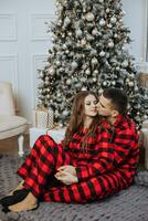 une magnifique Jeune fille dans rouge pyjamas des stands sur le rebord de fenêtre et met sur une Noël arbre. Noël concept. beaucoup cadeaux dans papier emballages en dessous de le Noël arbre photo