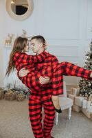 une magnifique Jeune fille dans rouge pyjamas des stands sur le rebord de fenêtre et met sur une Noël arbre. Noël concept. beaucoup cadeaux dans papier emballages en dessous de le Noël arbre photo