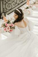 portrait de une Jeune fille de le dos, avec magnifique cheveux et maquillage dans une luxueux mariage robe sur le pas de une restaurant près Rose fleurs. verticale photo. photo