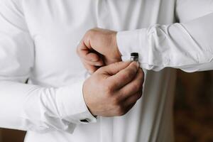 un adulte homme attache le manche de une blanc chemise, en train de préparer le jeune marié pour le mariage la cérémonie photo